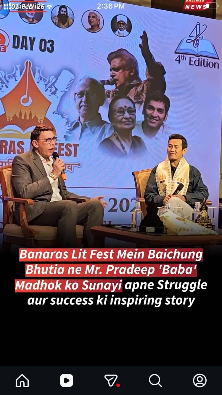 Two men seated in armchairs on a festival stage talk into microphones in front of a large printed backdrop.