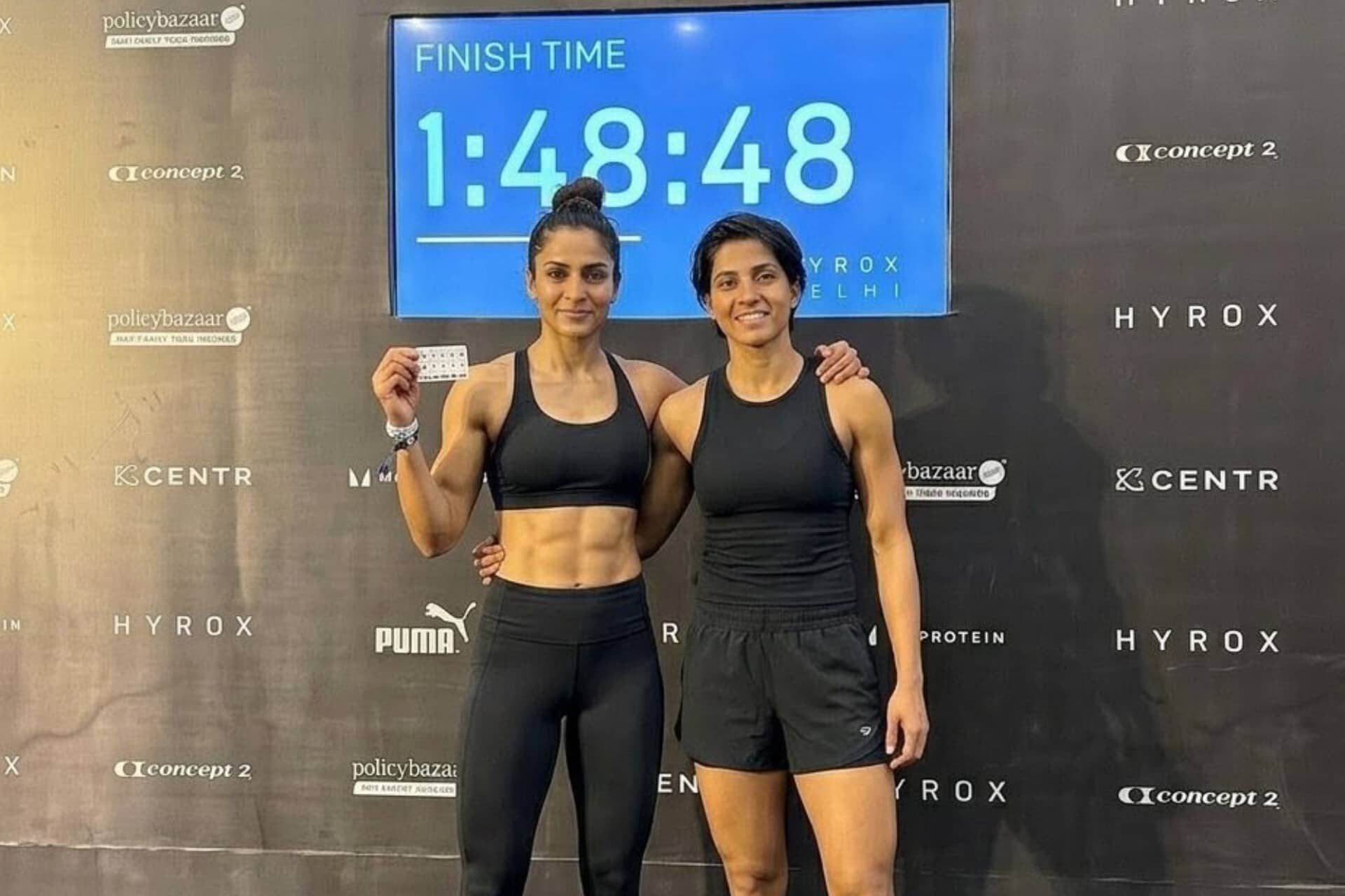 Two women in black athletic wear pose in front of a race finish-time screen showing 1:48:48.