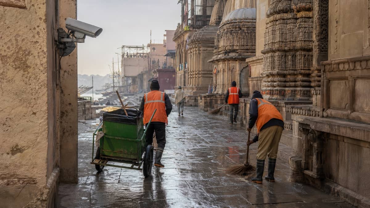 Sanitation workers and a waste cart in a narrow old Varanasi lane under CCTV