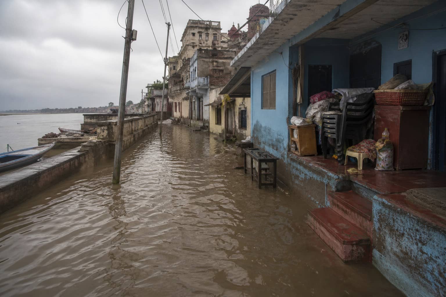 Illustrative flooded residential lane beside a monsoon river embankment with waterline marks on house walls