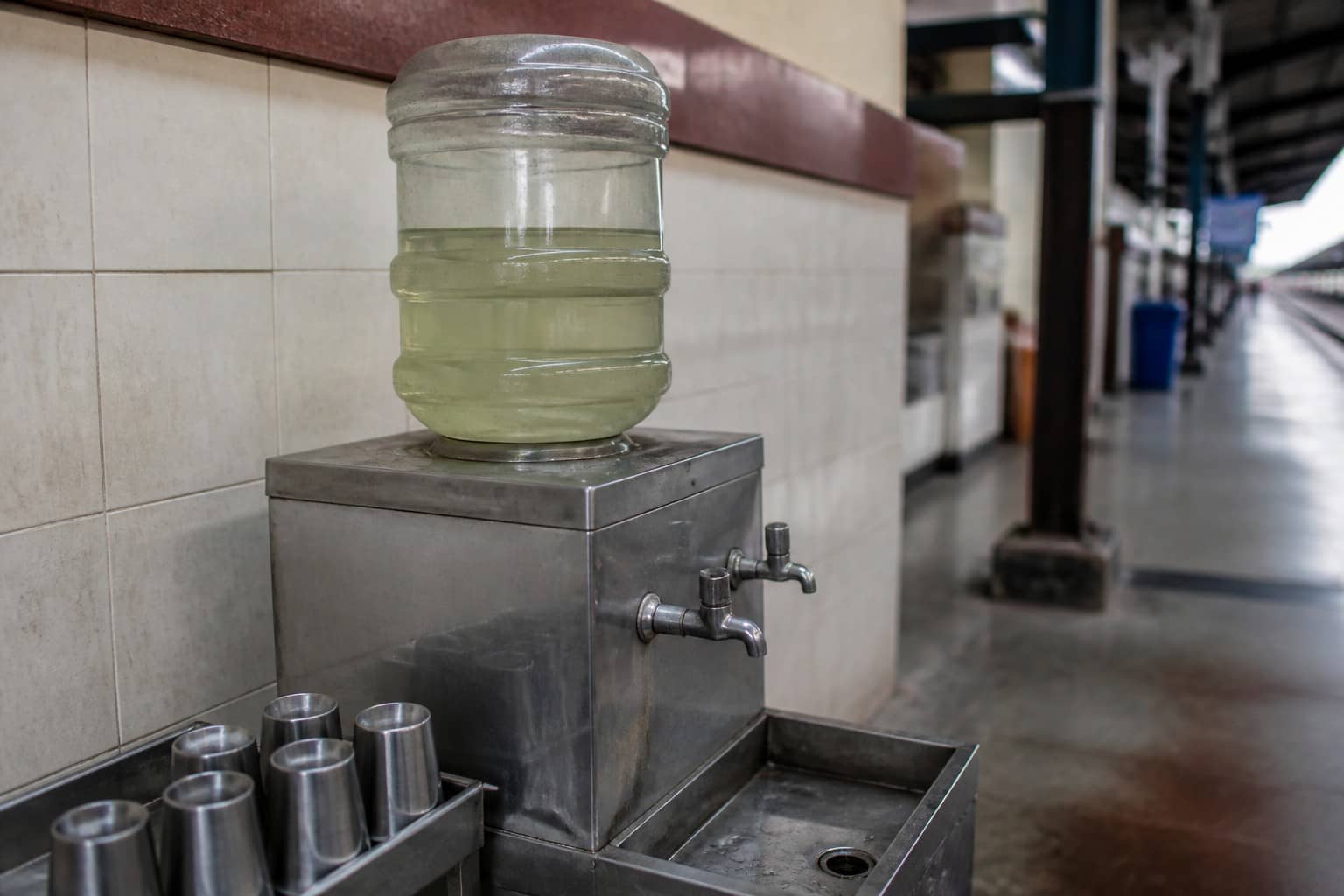 Illustrative railway-platform drinking-water booth with rusted tap and algae stain motif