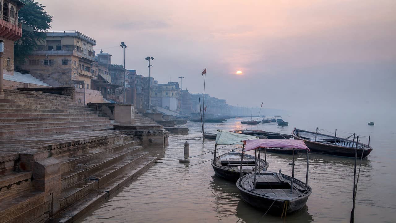 Illustrative photorealistic view of a swollen monsoon river covering stone ghat steps with mist and distant temple silhouettes along the Varanasi riverfront