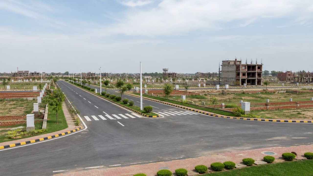 Illustrative photorealistic view of a planned residential layout with green belts, wide internal roads and distant low-rise housing under clear monsoon-season light