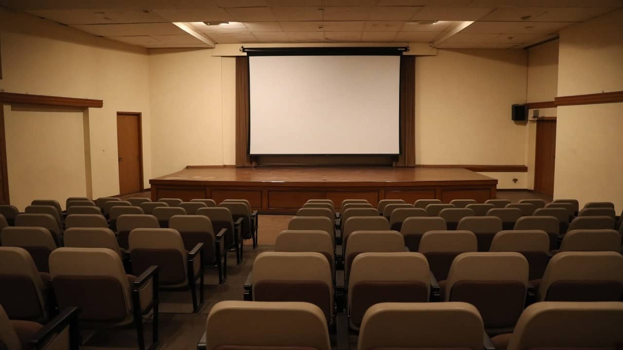 Photorealistic empty college auditorium with beige and brown seats facing a blank white projection screen, no logos or readable signs
