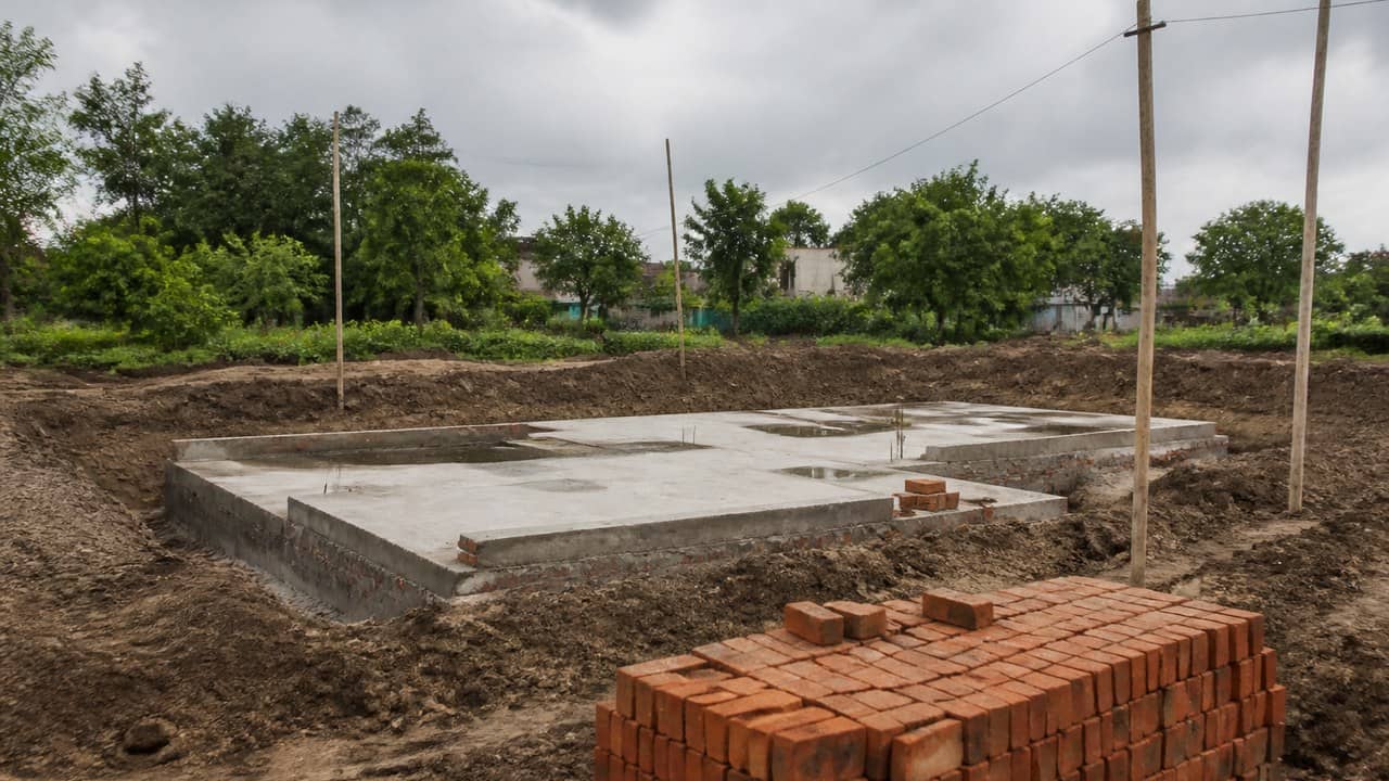Illustrative photorealistic empty civic construction site with a concrete foundation slab, stacked red bricks and overcast sky, no readable signs