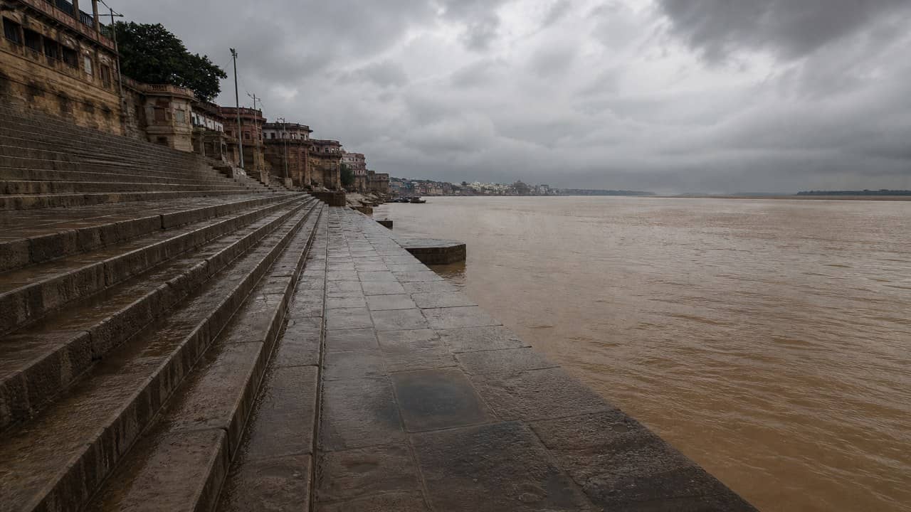 Photorealistic wet old brick rooftops and empty stone ghat steps on a north Indian river under a grey partly-cloudy monsoon sky, no people close, no readable signs