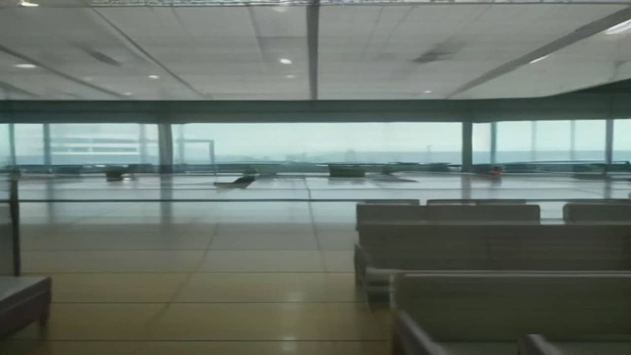 Empty modern airport terminal seating area with large windows looking onto a wet empty apron under overcast sky, no aircraft close, no people, no readable flight boards or logos
