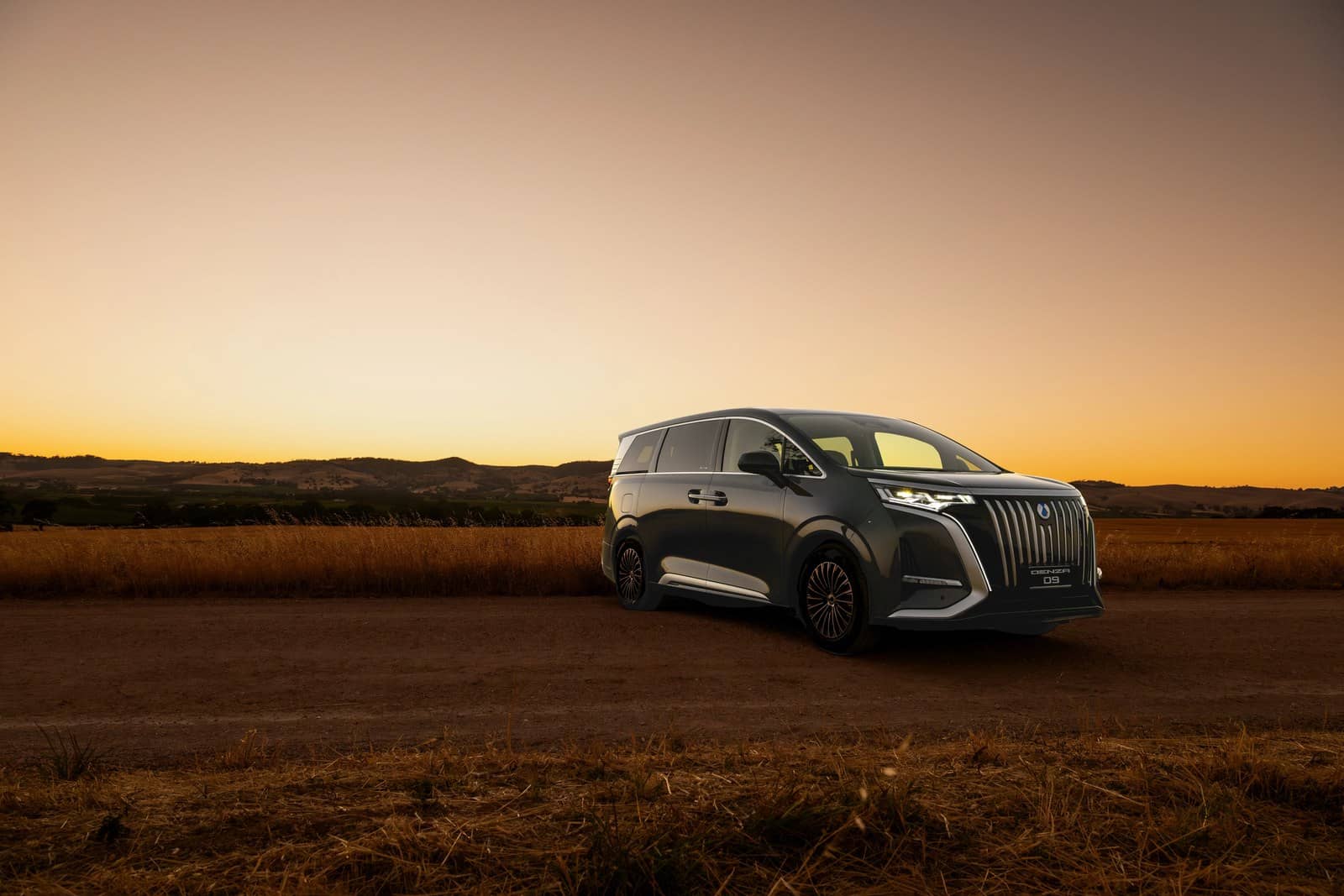 Dark grey Denza D9 luxury electric people mover seen from the front three-quarter angle with headlights lit, parked on a dirt road with golden grass hills and a sunset sky behind