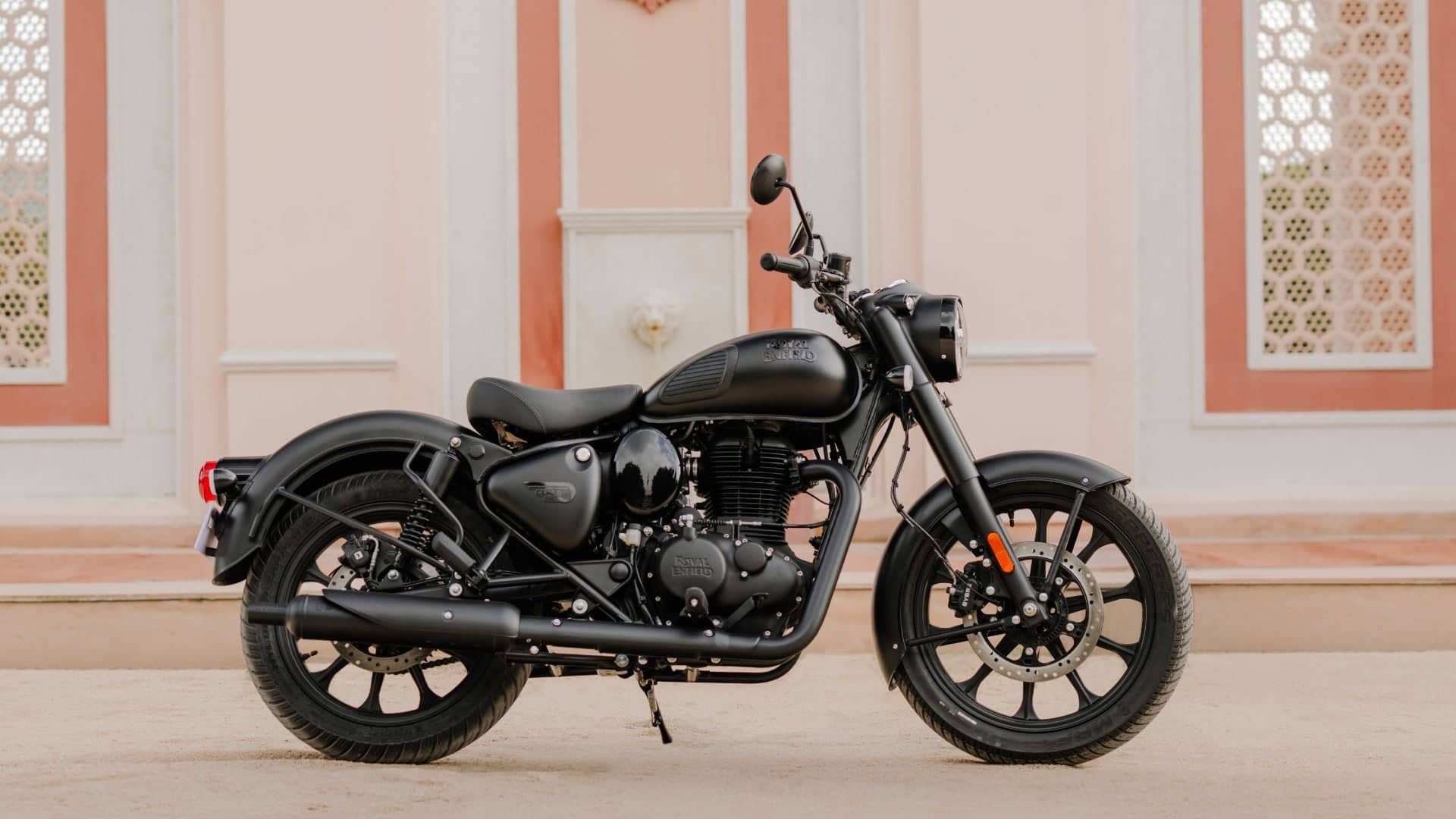 Matte black Royal Enfield Classic 350 with black spoked wheels parked side-on in front of a pink and white building