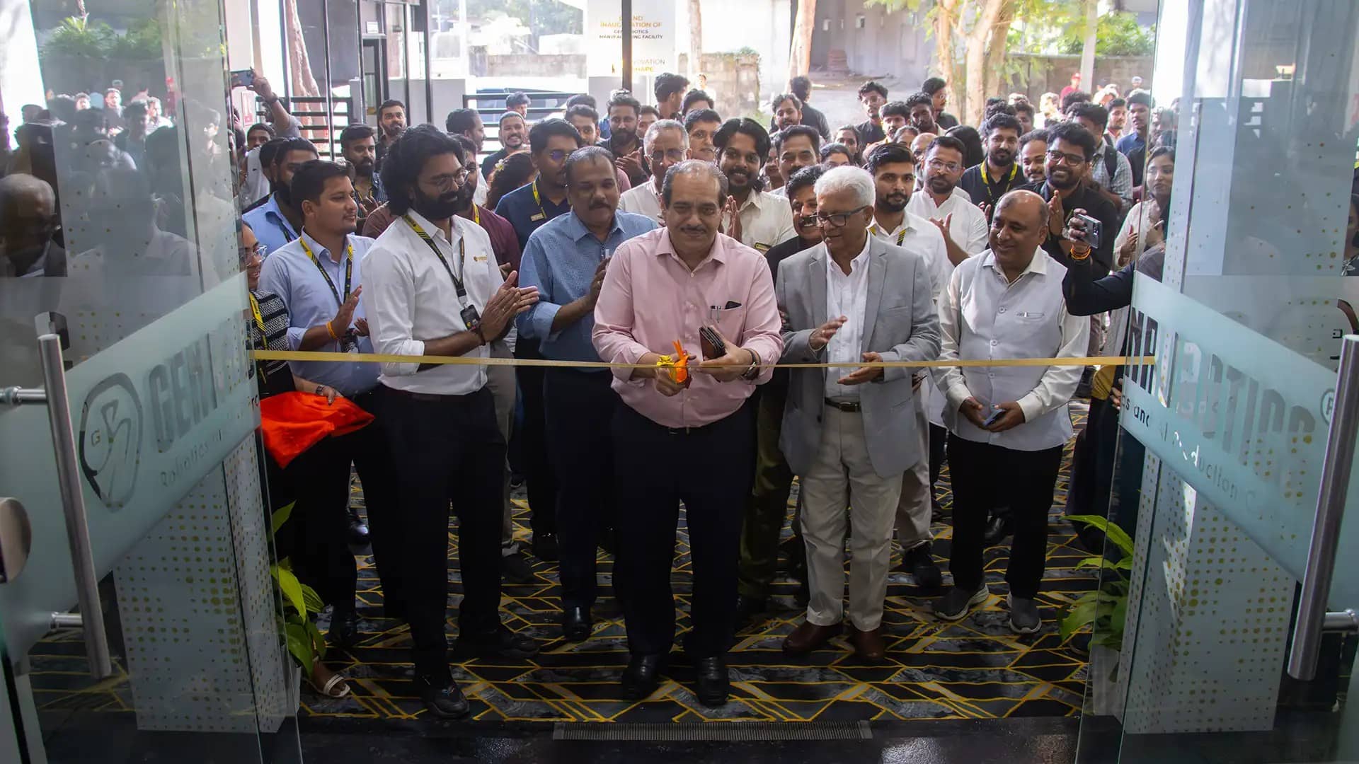 Officials and Genrobotics staff gather at a glass entrance as a man in a pink shirt cuts a yellow ribbon to open the facility, with a crowd clapping behind