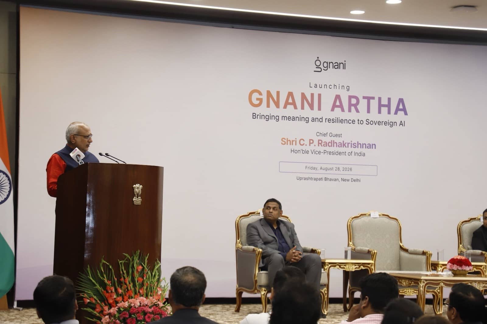 A man in a dark sleeveless jacket speaks at a wooden podium on a stage with a Gnani Artha launch backdrop, an Indian flag and armchairs, as guests watch