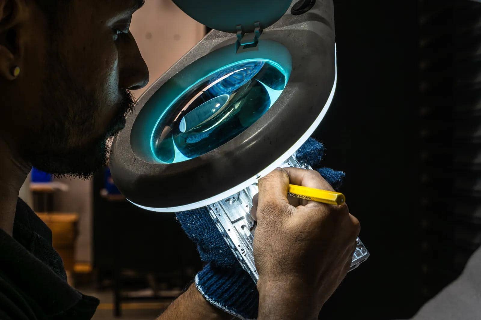 A worker looks through an illuminated magnifying lamp while marking a machined aluminium part with a yellow pencil