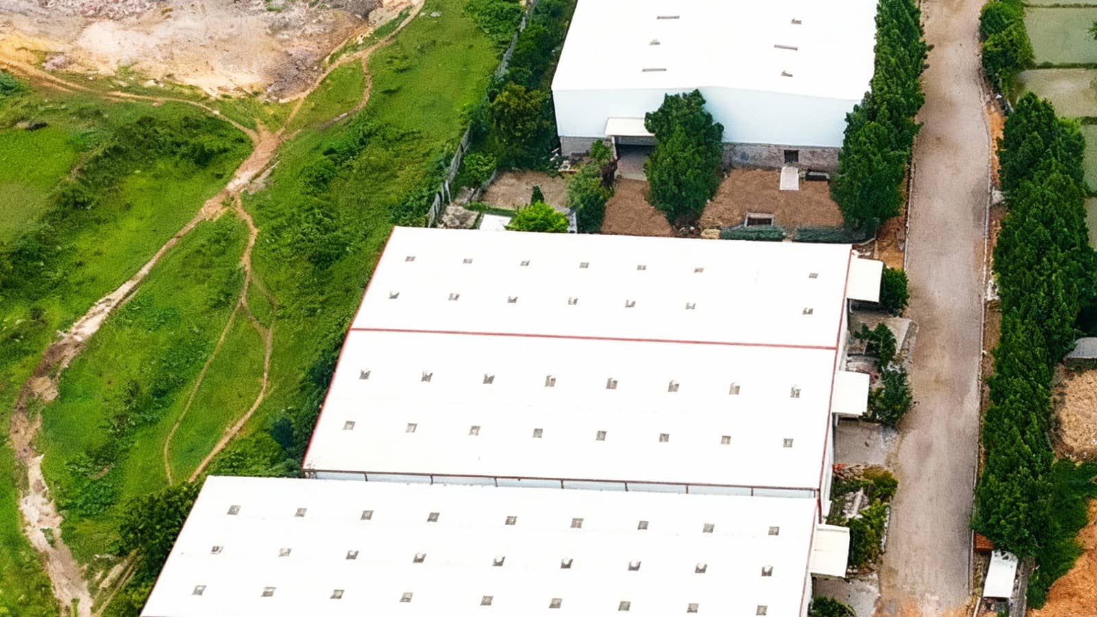 An aerial view of three large white factory sheds with small roof windows, surrounded by green trees and fields, at an AKSI Aerospace Group drone facility