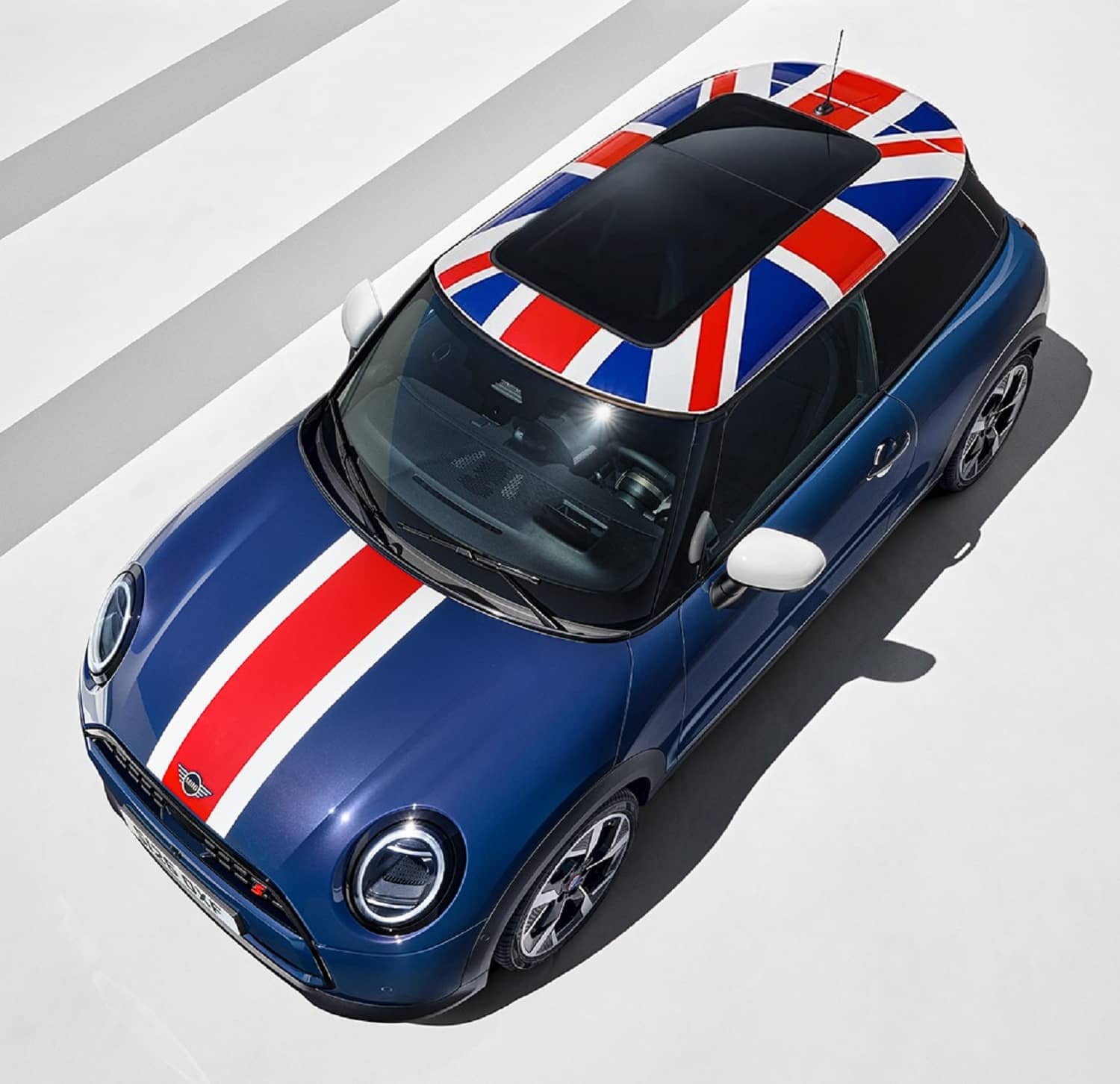 Overhead front three-quarter view of a blue MINI Cooper Oxford Edition with Union Jack roof graphics, red and white bonnet stripes and white mirrors