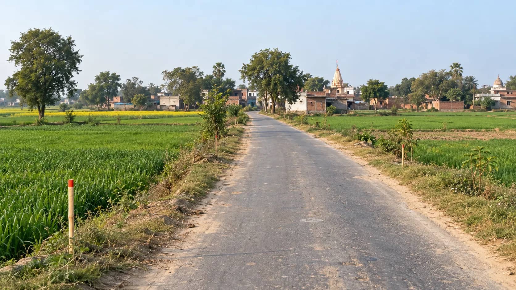 A narrow rural road runs between green fields towards a village, with survey stakes along the verge.