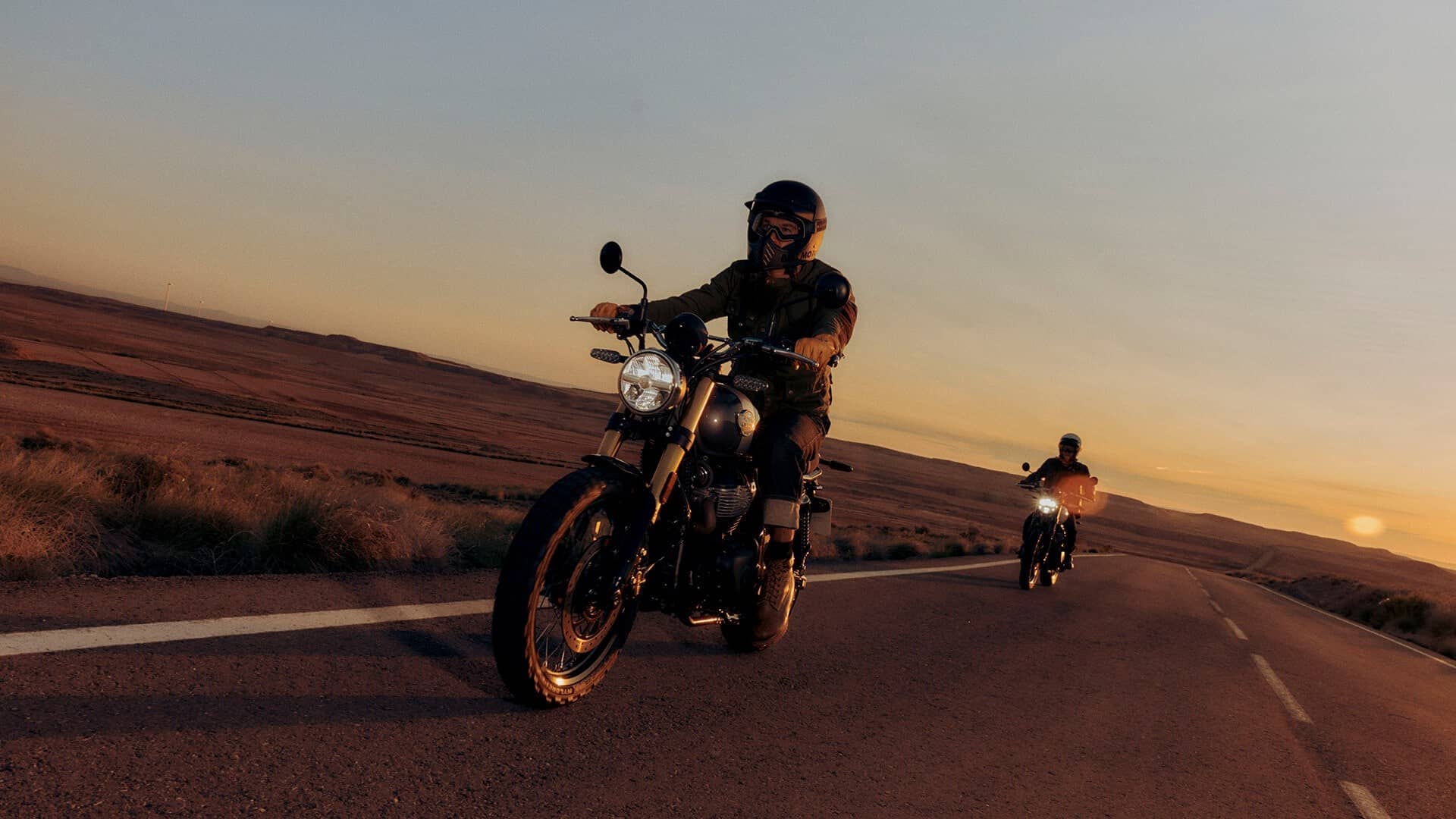 A rider on a Royal Enfield Bear 650 scrambler on an open road at dusk with a second rider in the distance