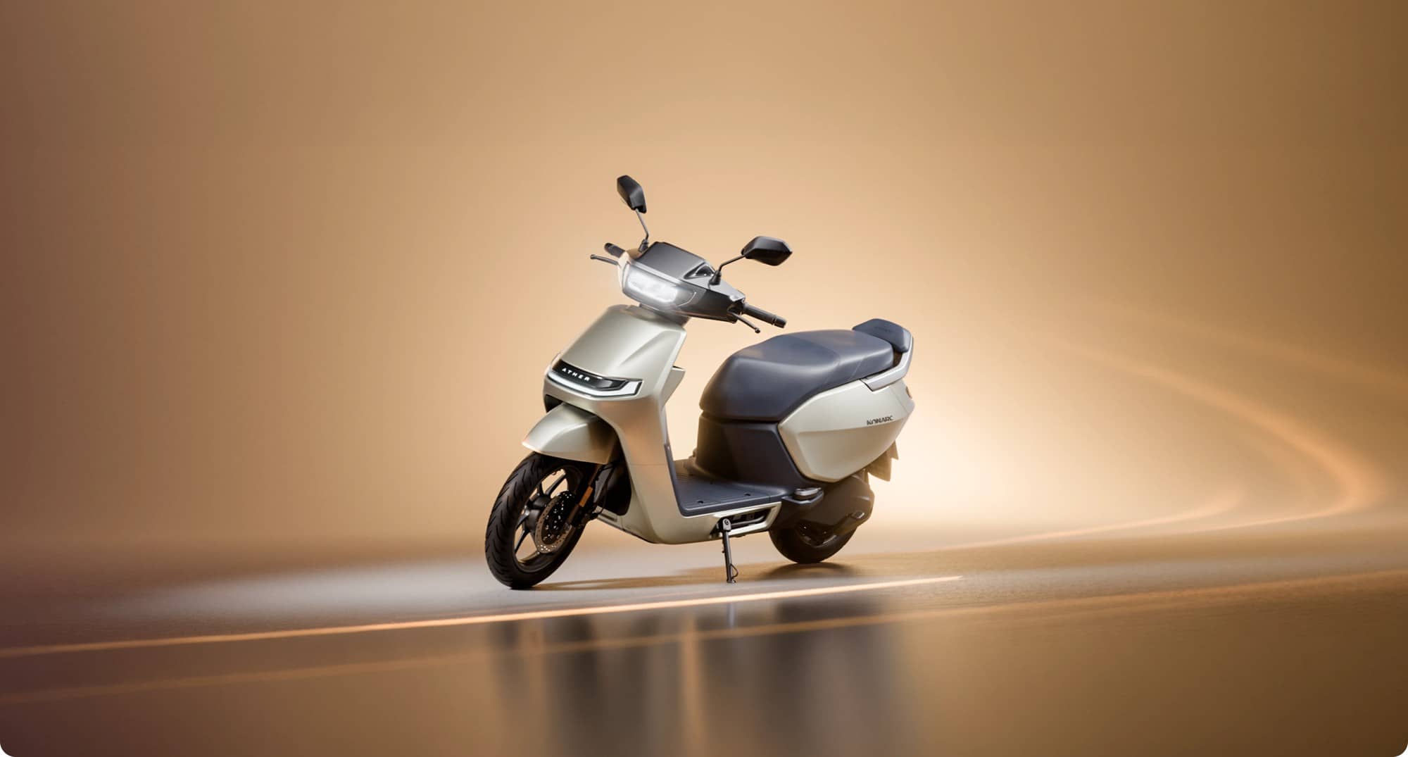 A silver Ather Konarc electric scooter with a dark seat standing on a warm brown studio floor with soft light trails