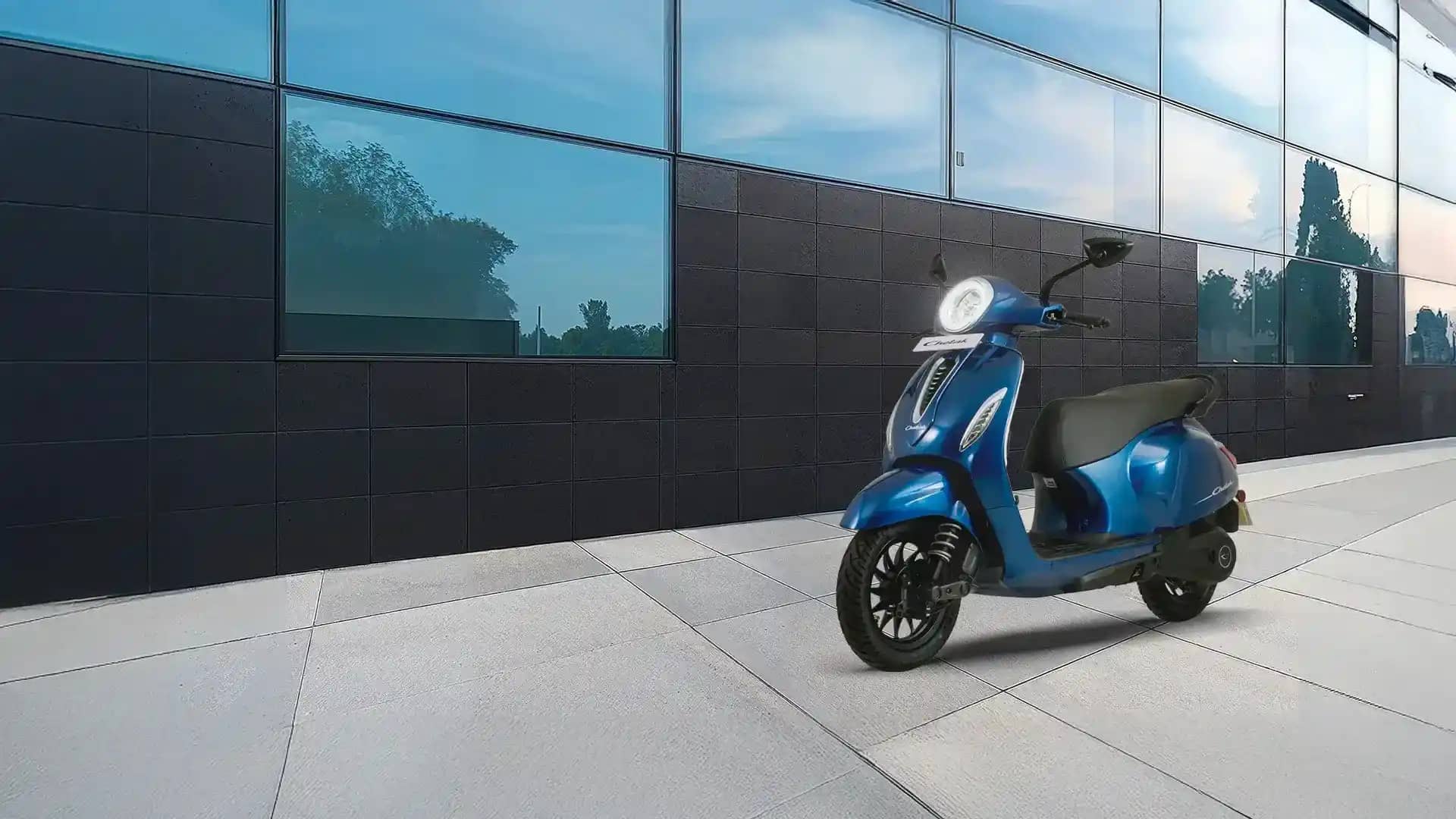 A blue Bajaj Chetak electric scooter with a round LED headlamp parked on a paved walkway beside a dark glass-fronted building