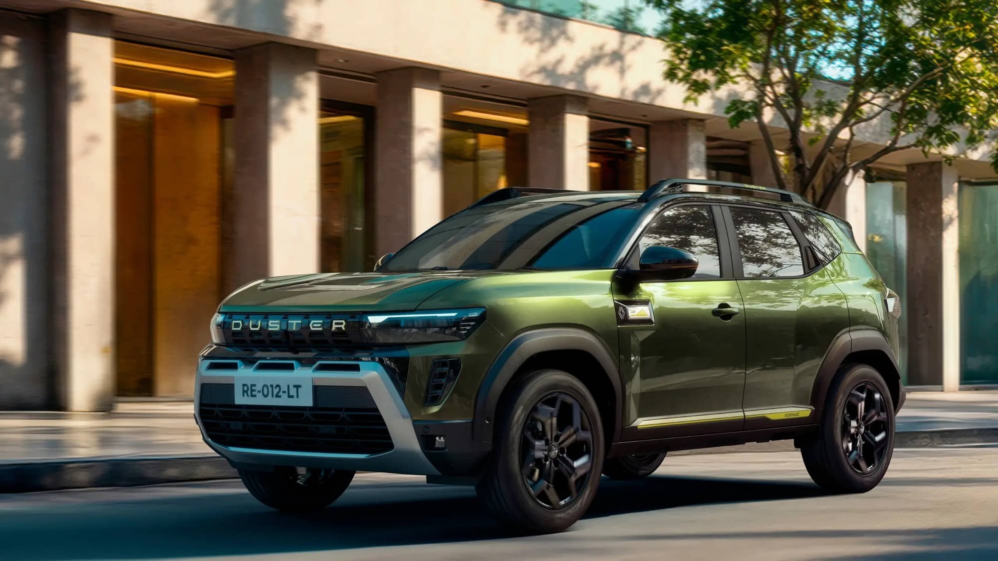 A green Renault Duster SUV seen from the front three-quarter angle driving past a building with a tree in the background