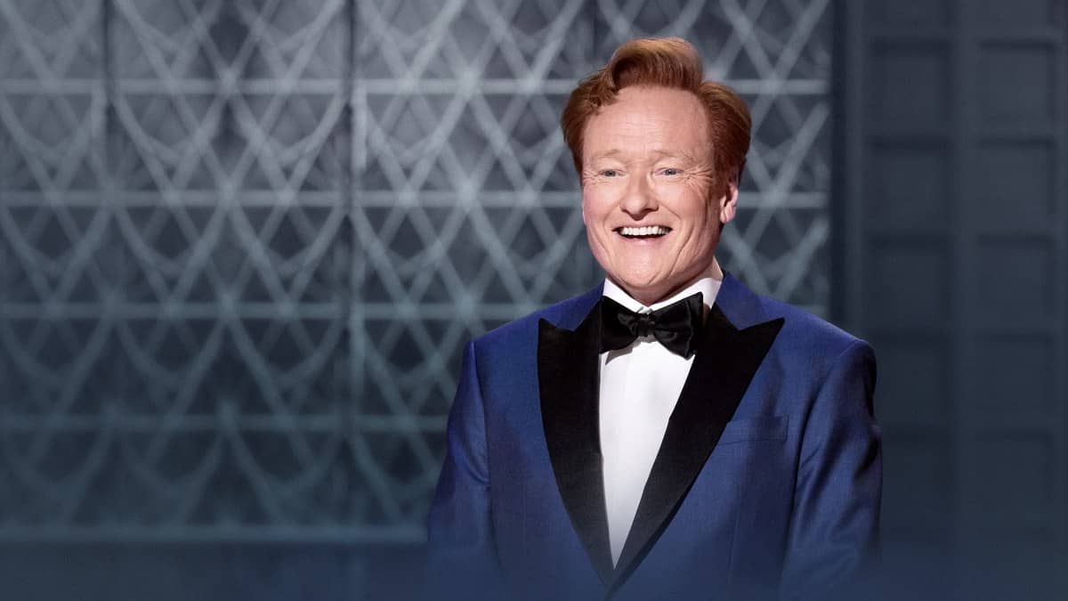 Conan O'Brien smiles in a blue tuxedo against a geometric backdrop in an Academy publicity photo.